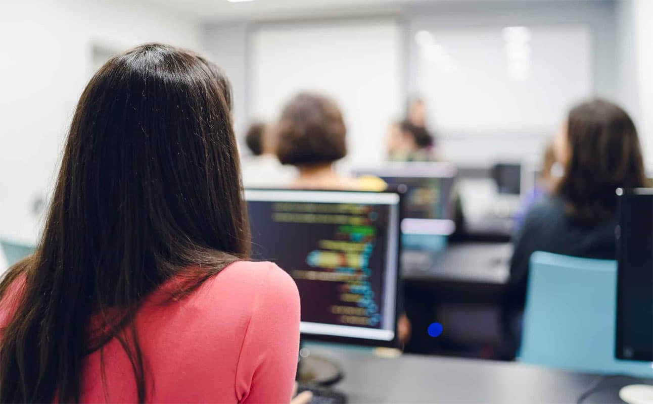 In einem Klassenzimmer des Lernstudios Barbarossa sitzt eine Gruppe von Menschen an Tischen mit Computern und konzentriert sich auf einen Code, der auf ihren Monitoren angezeigt wird.