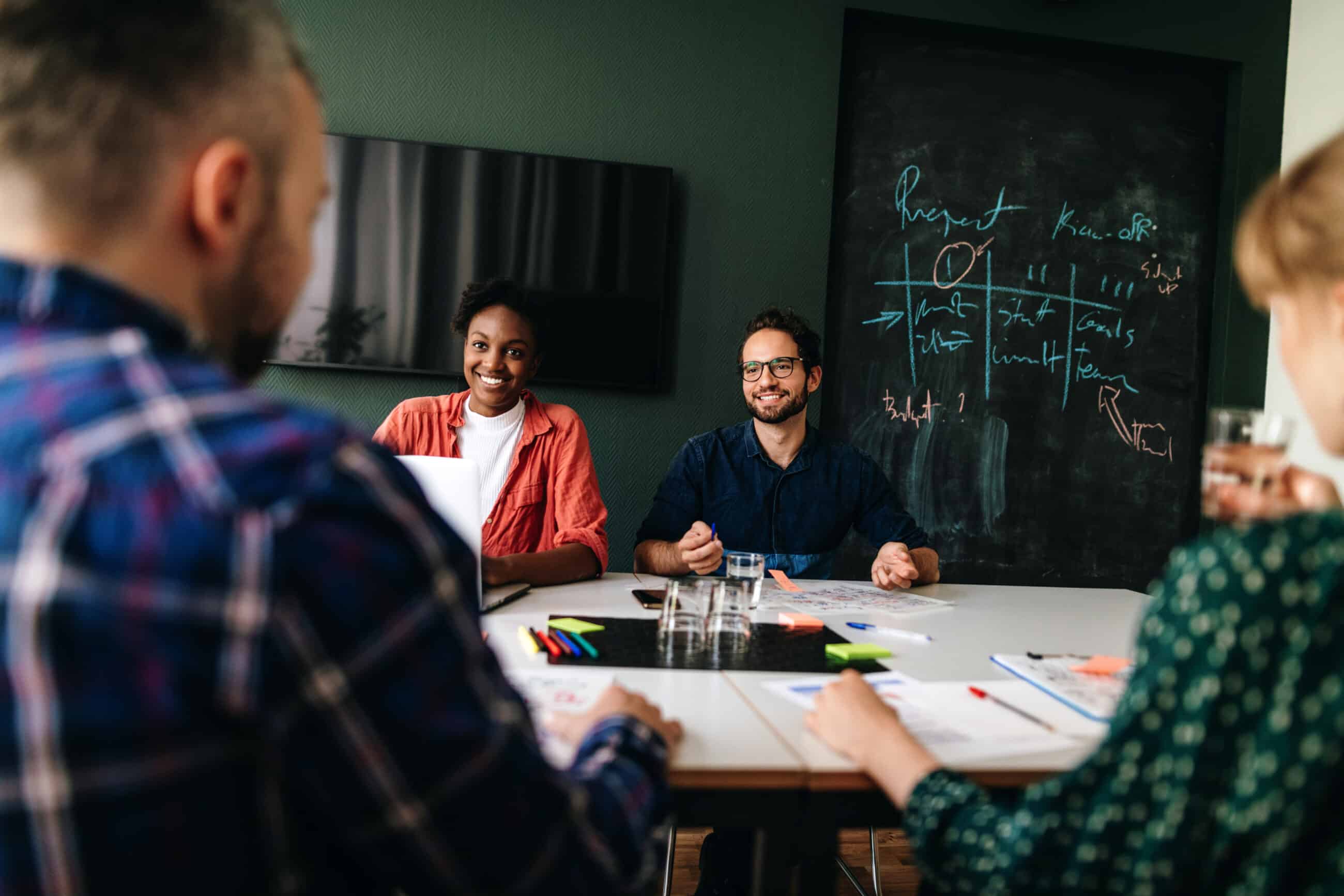 Vier Personen sitzen um einen Tisch in einem Besprechungsraum des Lernstudios Barbarossa und besprechen ihre Arbeit. Auf dem Tisch liegen Papiere, Stifte und Gläser, im Hintergrund ist eine Kreidetafel mit Diagrammen zu sehen.