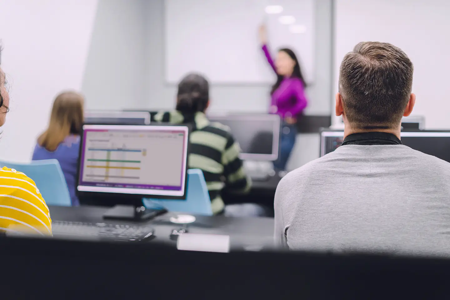Während eines Computerkurses sitzen Menschen an Computerarbeitsplätzen in einem Klassenzimmer, während eine Frau vorne steht und auf ein Whiteboard zeigt, während sie den Unterricht leitet.