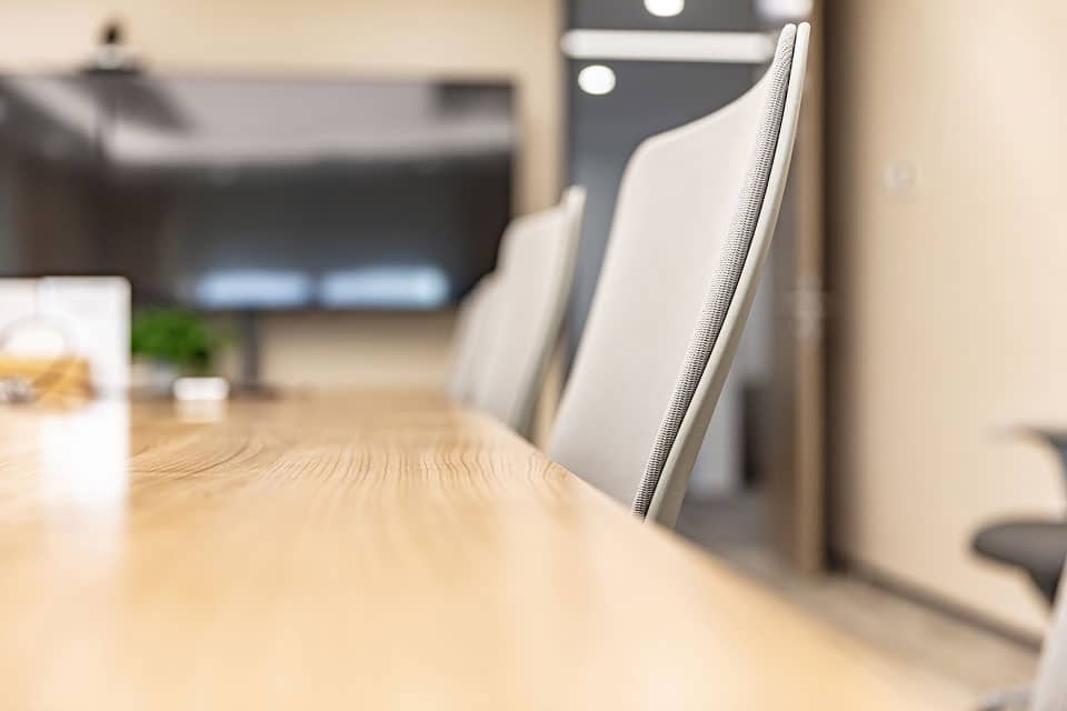 Nahaufnahme von modernen Bürostühlen, die an einem hölzernen Konferenztisch in einem Besprechungsraum aufgereiht sind, ideal für einen Computerkurs, mit einem großen Bildschirm im Hintergrund.