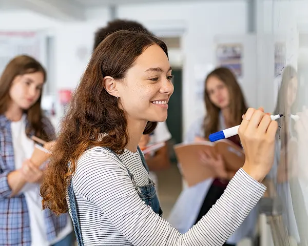 Junge Frau, die mit einem blauen Marker auf eine Tafel schreibt, während drei andere Schüler im Hintergrund zusehen.