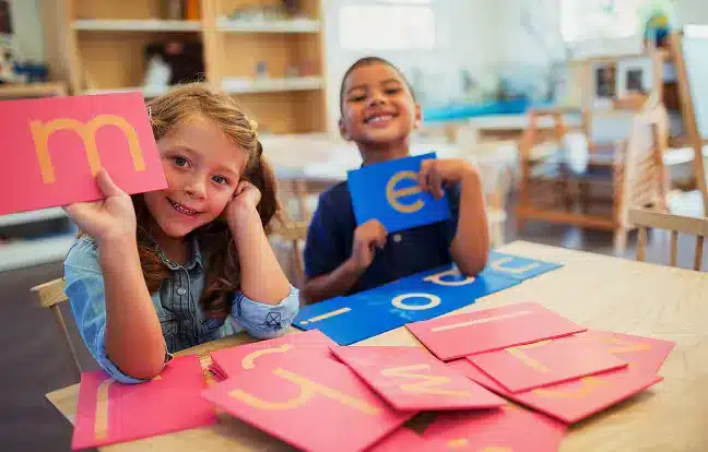 Zwei Kinder sitzen an einem Tisch in einem Klassenzimmer der Sprachschule, lächeln und halten Buchstabenkarten hoch. Weitere bunte Buchstabenkarten sind auf dem Tisch vor ihnen ausgebreitet.